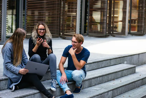 young adults sitting on steps smiling, 3 of them, one holding a laptop